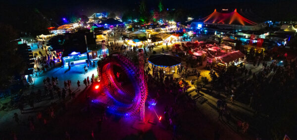 Aerial view of a bustling nighttime festival with colorful lighting, featuring rides, food stalls, and crowds of people enjoying the event.