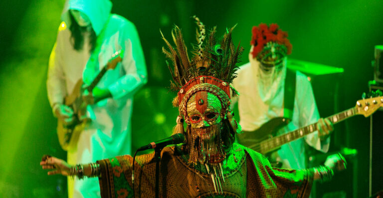 Performers in elaborate costumes and masks playing instruments on a stage with green lighting.