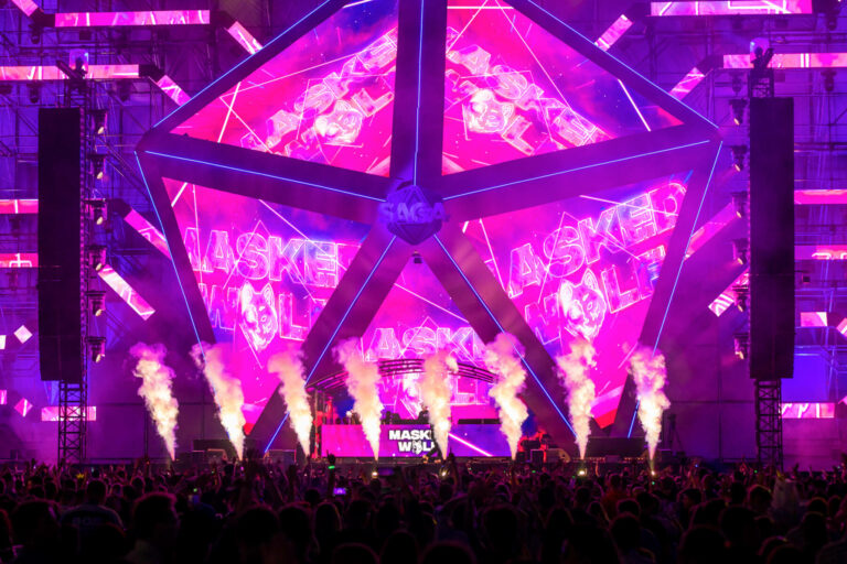 A vibrant electronic dance music concert with a large crowd, featuring a stage with dynamic purple and blue lighting, geometric shapes, and plumes of smoke.