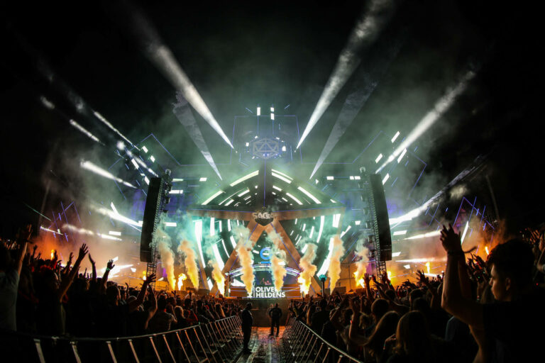 Crowd of people with raised hands at a night-time outdoor music festival with a lit stage, pyrotechnics, and laser light show.