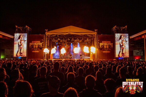 Crowd of people watching a live band perform on an outdoor stage with pyrotechnics at night, flanked by large screens displaying a guitarist, at Dynamo Metalfest in Eindhoven.