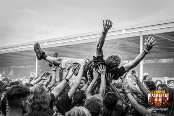 Black and white image of a person crowd surfing at a music festival, with many hands supporting them. The background shows a large tent structure and a crowd. The logo for 