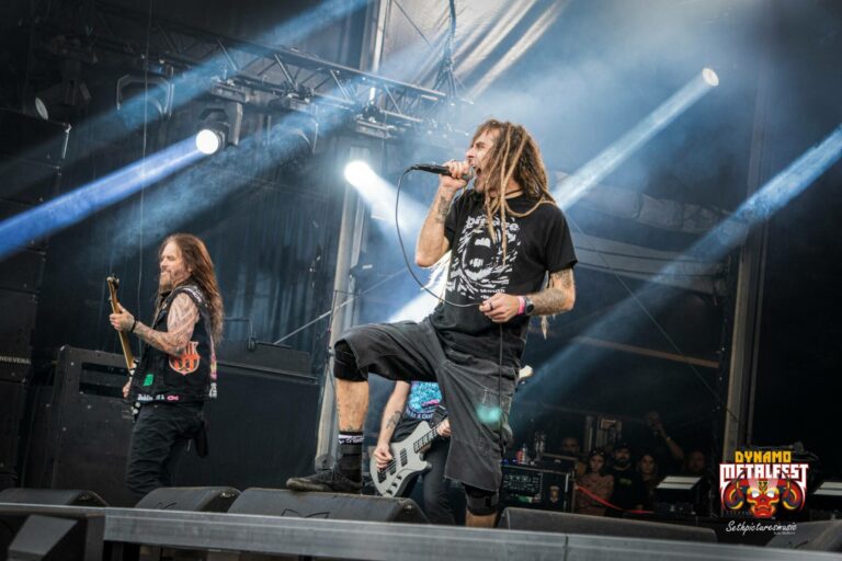 Two band members performing at Dynamo Metalfest, with the guitarist on the left and the vocalist on the right, both with long hair, under blue stage lighting with a smoke effect.