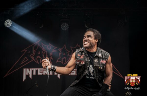 A male vocalist on stage at a metal festival, passionately singing into a microphone, with band logos in the background and stage lights shining down.