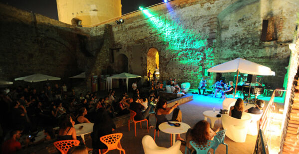 An outdoor evening event with people sitting and standing in a courtyard, listening to a live band performing under a green illuminated stone wall and a tower.
