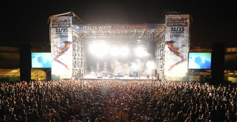 A large crowd at the Italia Wave Love Festival concert with stage lights, banners showing the event name, and a live band performing at night.