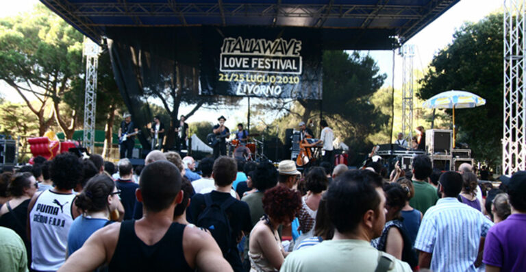 An outdoor music festival scene with a crowd of people watching a live band perform on stage, with a banner above the stage reading 