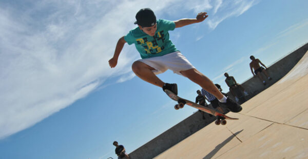A young skateboarder executing a trick at a skatepark with onlookers in the background under a clear blue sky.