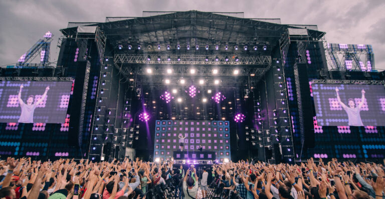 A large crowd at an outdoor music festival cheering with hands raised in front of a stage with multiple screens displaying a performer and purple stage lights.