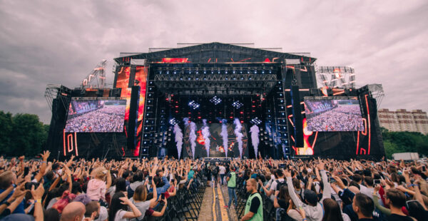 Crowd of people enjoying an outdoor concert with hands raised in front of a large stage with bright screens and lighting equipment.