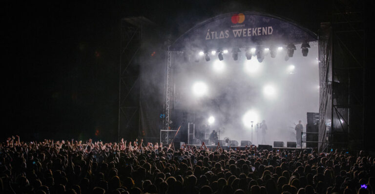 Outdoor nighttime concert scene with a large crowd of people watching a performance on a stage illuminated by bright lights, featuring a banner with "ATLAS WEEKEND" written on it.