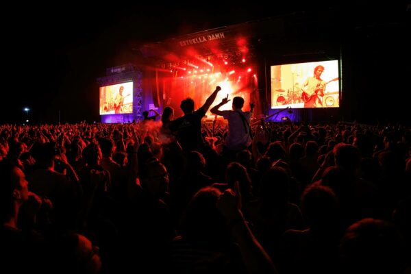 A large crowd at an outdoor music festival at night, illuminated by red stage lights and flanked by large screens showing musicians performing.
