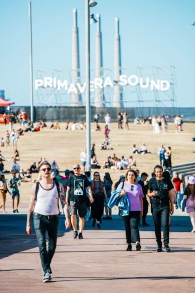 People walking and sitting outside at the Primavera Sound festival with large lettering of the event's name in the background and industrial structures against a clear blue sky.
