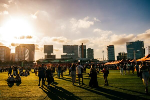 A group of people at an outdoor event with artificial turf, with the sun setting in the sky and city buildings in the background.
