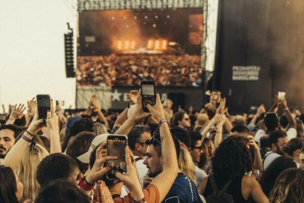 A crowd of concert-goers raising their hands and smartphones to capture the event, with a large stage screen visible in the background.