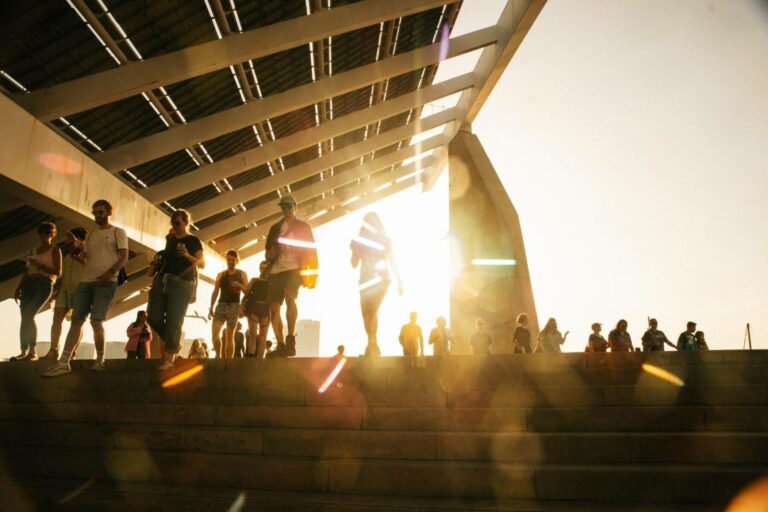 People walking down steps outdoors, with the sun creating silhouettes and glare, under a large structure's shadow.