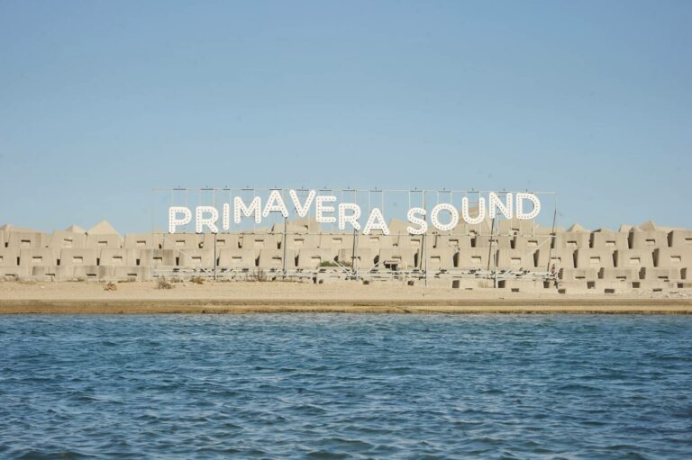 Large white letters spelling "PRIMAVERA SOUND" installed on poles in front of a line of identical, uniform concrete buildings near a body of water under a clear blue sky.