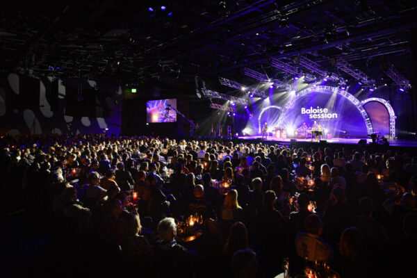 A crowded concert hall with candle-lit tables, spectators watching a live performance on stage under blue and purple lighting, with the words 