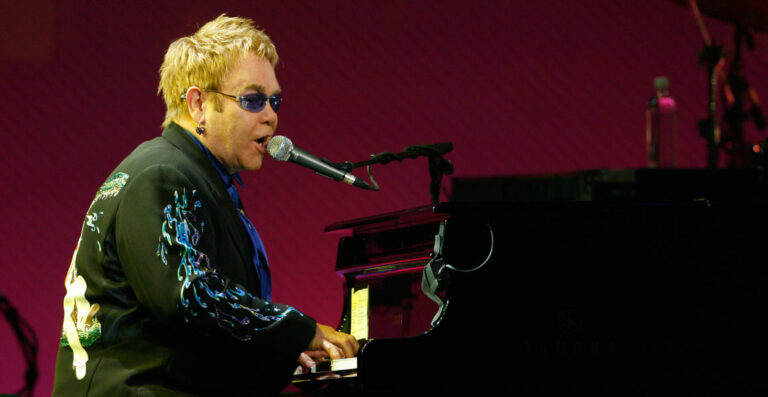 A person with sunglasses and a decorative jacket playing a grand piano and singing into a microphone onstage with a purple backdrop.