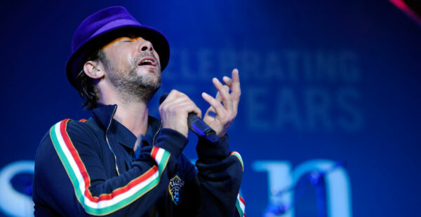 A man wearing a purple hat and a black jacket with colorful stripes on the sleeves passionately singing into a microphone with a blue stage background that includes text 