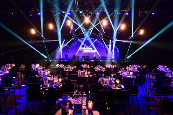 A concert hall set up for a formal event with round tables, candles, and wine glasses, with a stage at the front displaying 'Baloise Session' in large lettering, illuminated by dramatic blue stage lights.