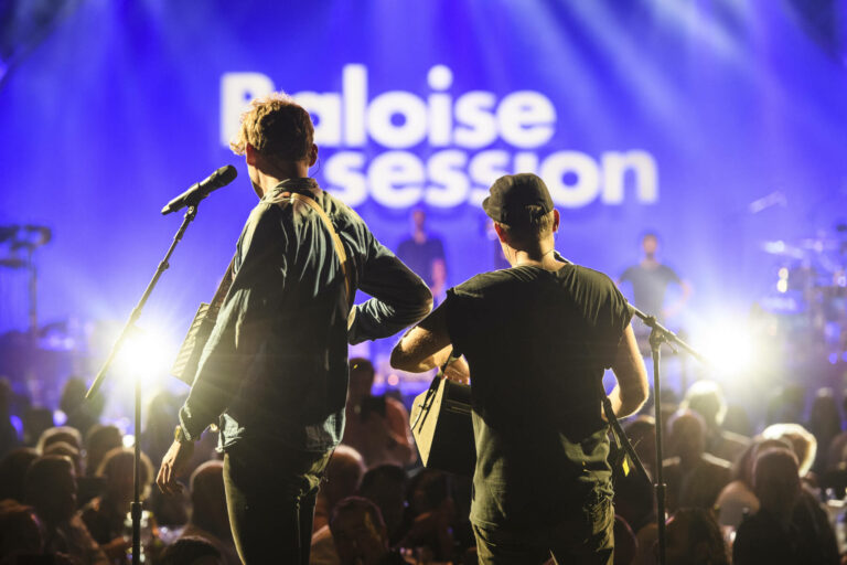 Two musicians are seen from behind performing on stage at a concert with the audience in the foreground and the words 