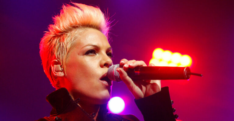 A person with a short, spiky hairstyle singing into a microphone with bright lights in the background.