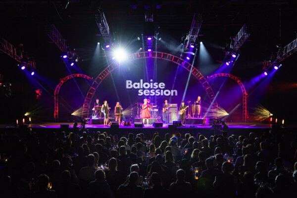 A live concert performance at the Baloise Session with an audience seated at tables in the foreground and a band performing on a lit stage with the event name in the background.