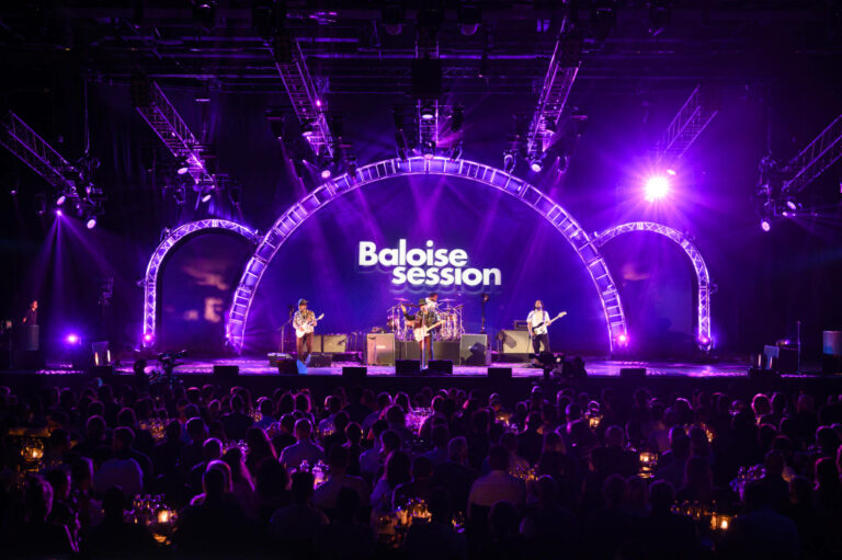 A band performing on stage at the 'Baloise Session' music festival with audience members seated at tables, stage lights, and a large screen with the festival's logo in the background.