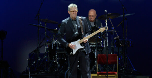 Two men on stage during a concert, one playing an electric guitar and the other playing drums in the background, both dressed in formal black attire with a dark blue backdrop.