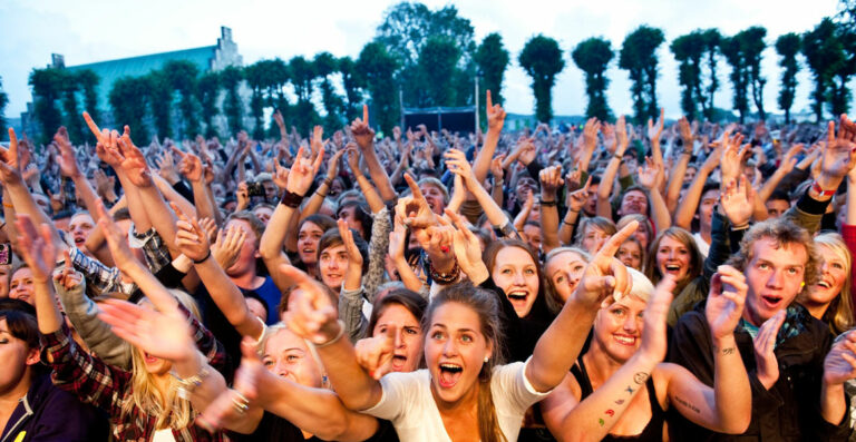 A large crowd of enthusiastic concert-goers cheering and raising their hands in the air, some making the 'rock on' hand gesture, during an outdoor music festival.
