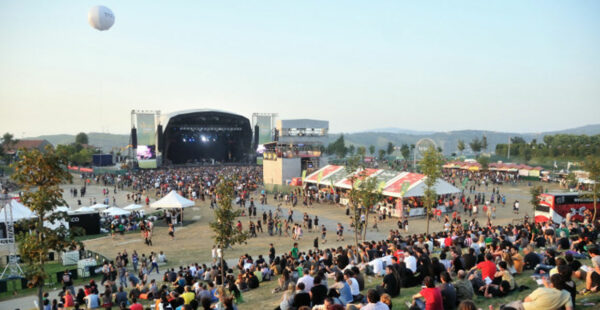 Outdoor music festival with a crowd of people, some seated and others standing, with a stage in the distance and food booths scattered around the venue.