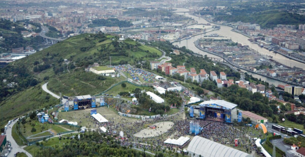 Aerial view of an outdoor music festival with multiple stages, crowds of people, and tents set up on a grassy area near a city with a river running through it.