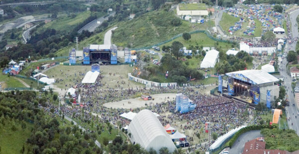 Aerial view of an outdoor music festival with large crowds, multiple stages, tents, and surrounding greenery.