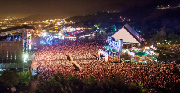Aerial night view of a crowded music festival with large stages and bright lights surrounded by trees.