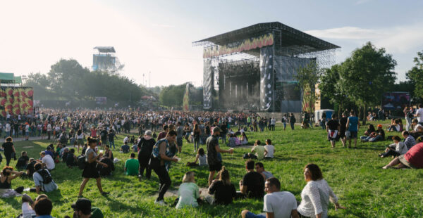 A large crowd of people gathered on a grassy hill at an outdoor concert with a stage in the background and a clear sky above.