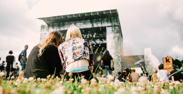 Two people sitting on the grass at an outdoor music festival with other attendees standing around and a stage in the background.