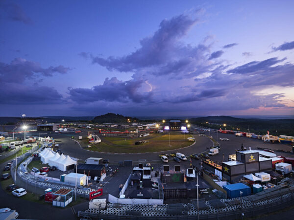 A twilight view over a race track complex with tents, trucks, and lighting, under a dramatic sky showing a hint of lightning in the distance.