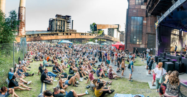 A panoramic view of an outdoor music festival with a large crowd of people sitting and standing in front of a stage with musicians performing, surrounded by industrial structures and clear blue skies.