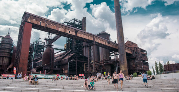 A wide-angle view of an old industrial steel plant with large rusted structures and pipes, with a group of people relaxing and walking on the stairs in the foreground under a partly cloudy sky.