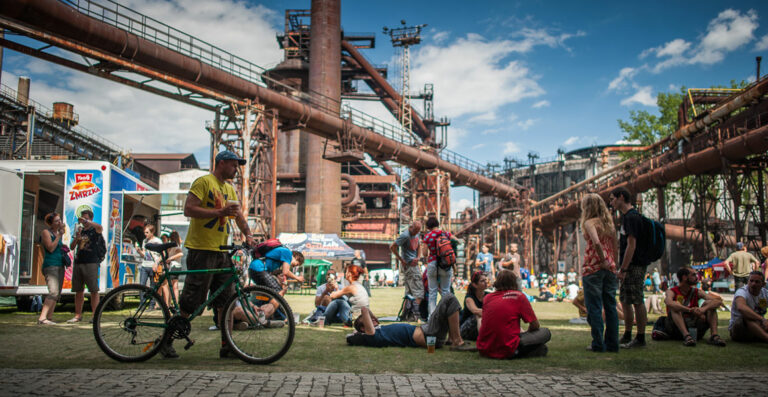 People socializing in a park with an industrial backdrop featuring large rusted steel structures and pipes, with a man in the foreground walking beside a bicycle.