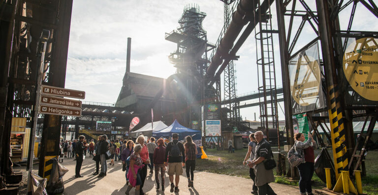 A sunny day at an outdoor event with people walking around industrial structures, including a large decommissioned blast furnace. Directional signs are in the foreground with multiple paths leading to various attractions, creating a bustling atmosphere.