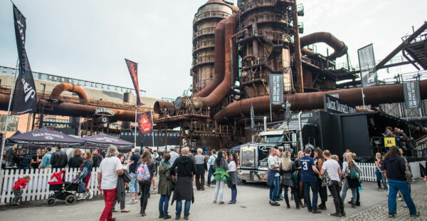 A bustling outdoor event with people gathered around food trucks and stands with banners under the rustic backdrop of an old industrial structure with large pipes and metallic towers.