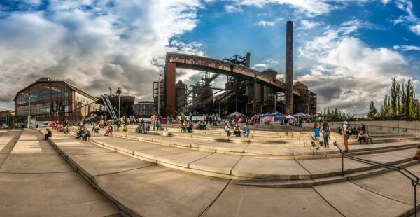 Panoramic view of a public gathering in an industrial park with historical steel structures, people milling about and seated on wide stone steps under a partly cloudy sky.