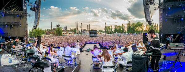 Panoramic view of an outdoor concert from the stage perspective, showing an orchestra in the foreground and a massive audience in the background, with industrial structures and a clear sky.