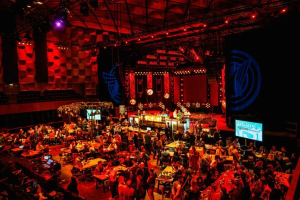 A vibrant indoor event space filled with attendees, featuring a stage with red lighting, tables with guests, and decorative plants under a ceiling with geometric paneling. A large blue logo is projected on the right wall.