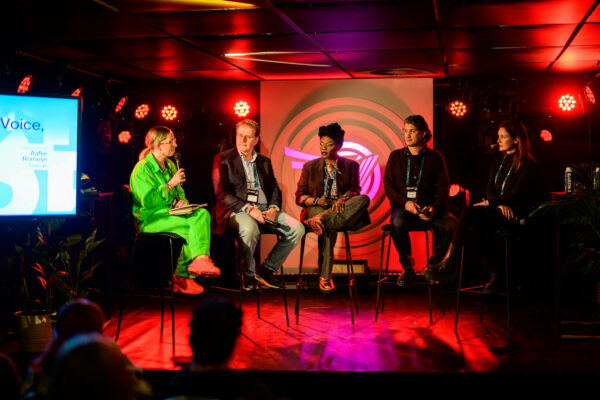 Four people sitting on stage for a panel discussion at a conference with colorful stage lighting and a presentation screen visible to the left.