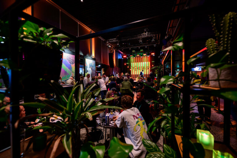 A lively indoor event space with people socializing at tables, vibrant lighting, and decorative plants in the foreground.
