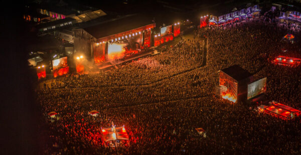 Aerial view of a large outdoor concert at night with dense crowds and several stages illuminated by red and orange lights.