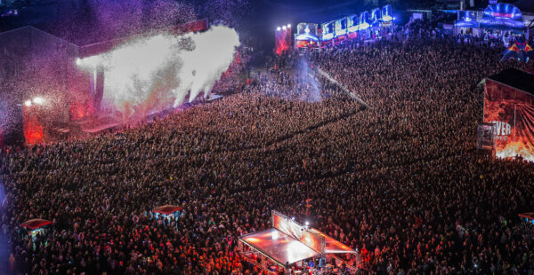 A vast crowd of people at an outdoor music festival at night, with a stage lit with red lights and white smoke effects.